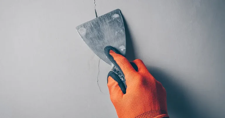 A hand in an orange glove holds a spatula to fix a crack in a wall.