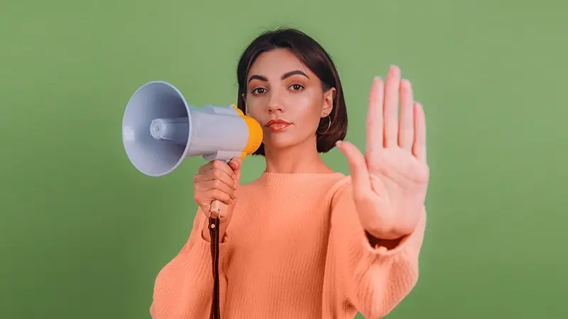 A woman on a green background holds a megaphone in one hand and makes a stop gesture with the other.