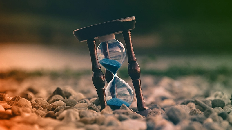An hourglass with blue sand, gently resting on gray and white stones, captures a subtle harmony between time and nature.