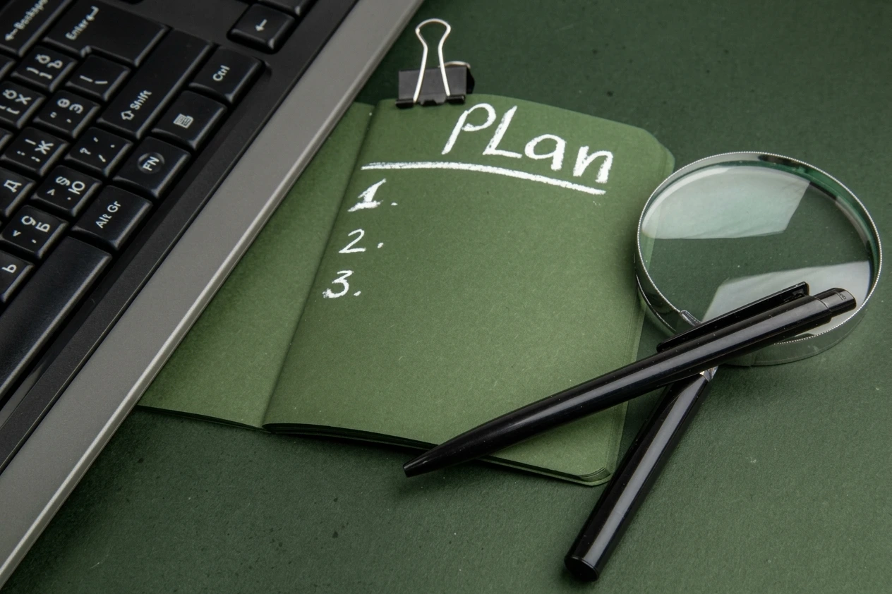 Carnet vert avec « Plan », loupe, stylos, clavier d'ordinateur.